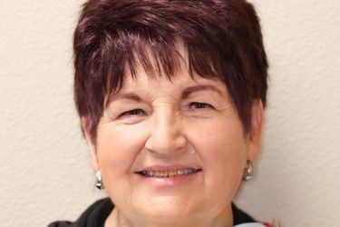 A smiling older woman with short, dark red hair and earrings, against a neutral background.