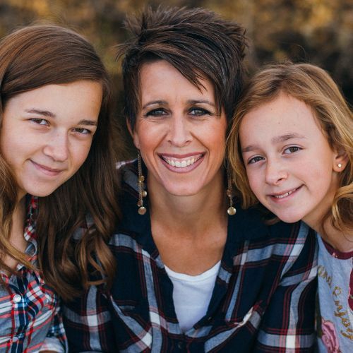Woman with two young girls, all smiling, wearing plaid shirts. Outdoors, soft lighting.