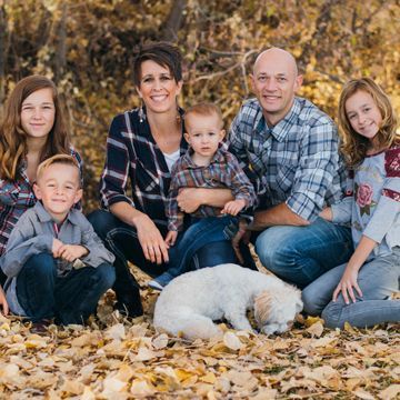 Family of seven, including a small dog, sitting in fallen autumn leaves. They smile for the camera, dressed in casual attire.