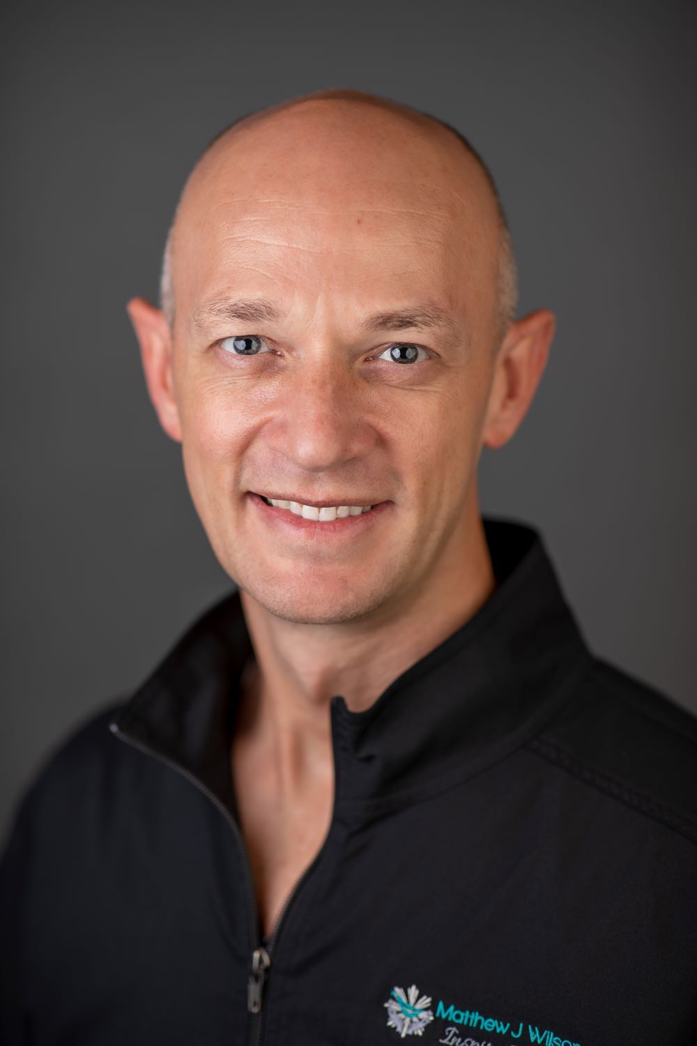 A smiling, fair-skinned man with a shaved head wearing a black jacket with a small logo against a gray background.
