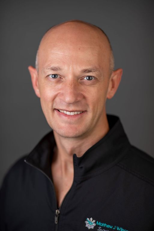 A smiling, fair-skinned man with a shaved head wearing a black jacket with a small logo against a gray background.