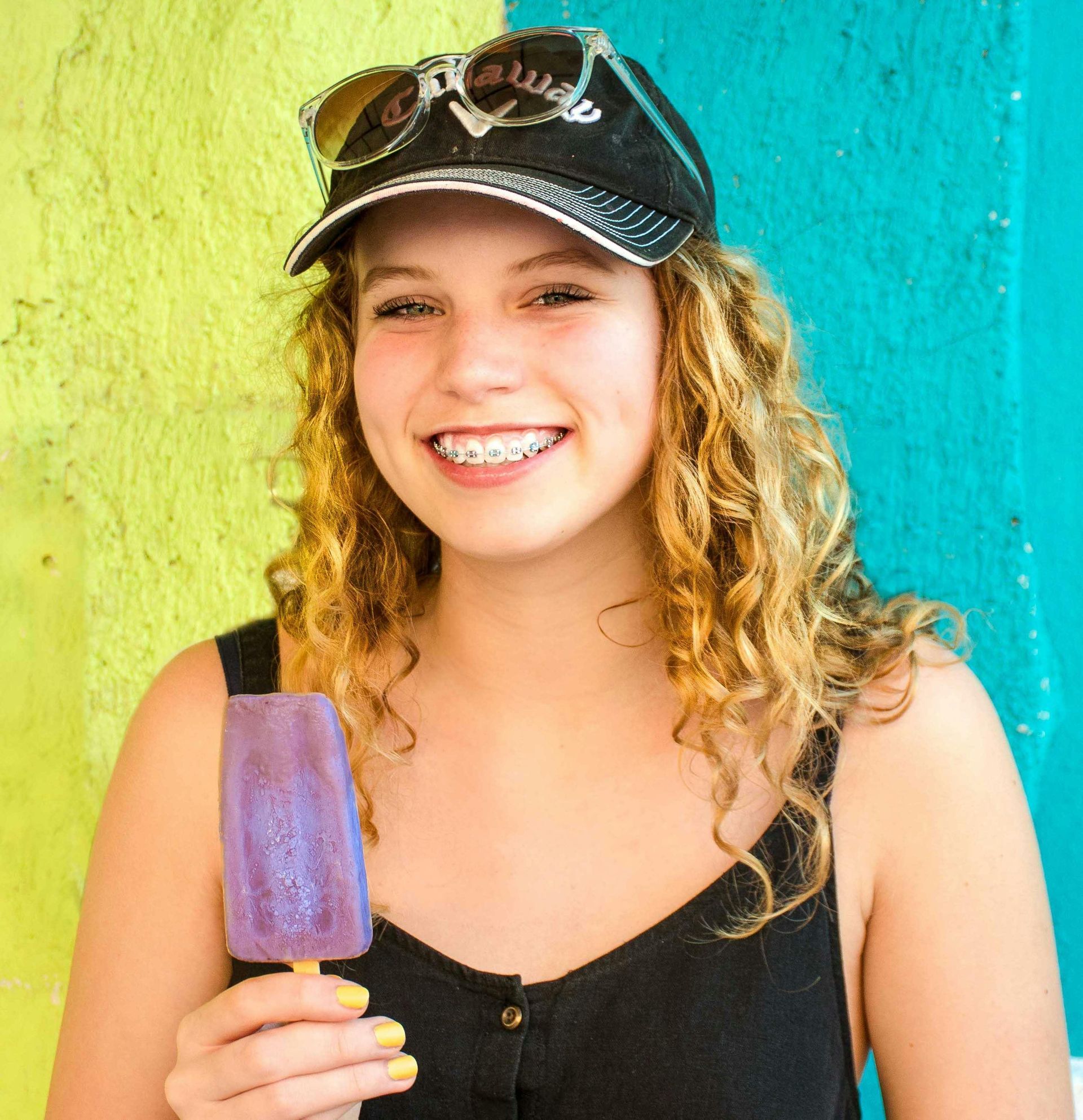 Young woman with curly hair and braces, wearing sunglasses on a cap, holding a purple popsicle in front of a colorful wall.