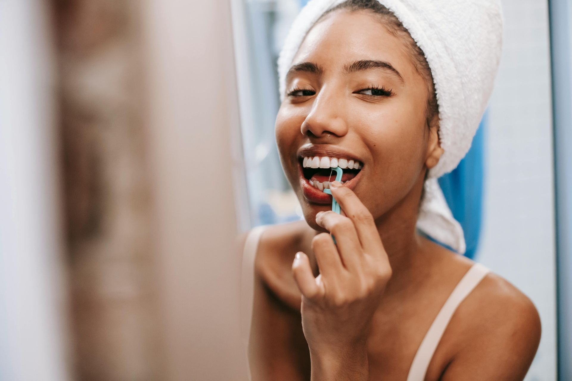 Woman with a towel on her head, smiling and cleaning her teeth with a small interdental brush in a bathroom setting.