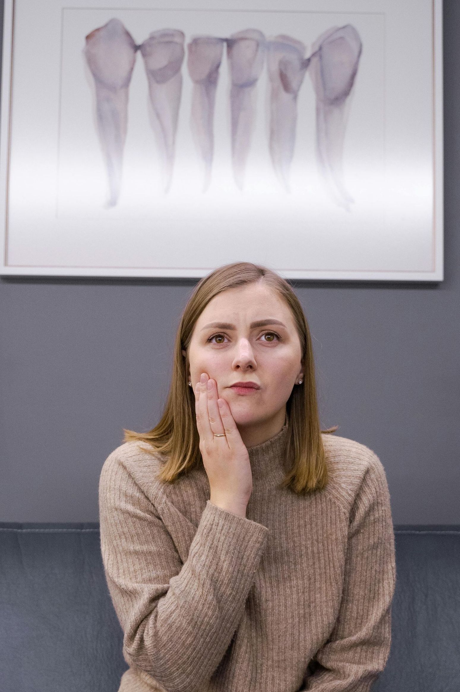 Woman in a beige sweater holding her hand to her jaw, looking pained. Above her hangs a framed artwork of tooth roots.