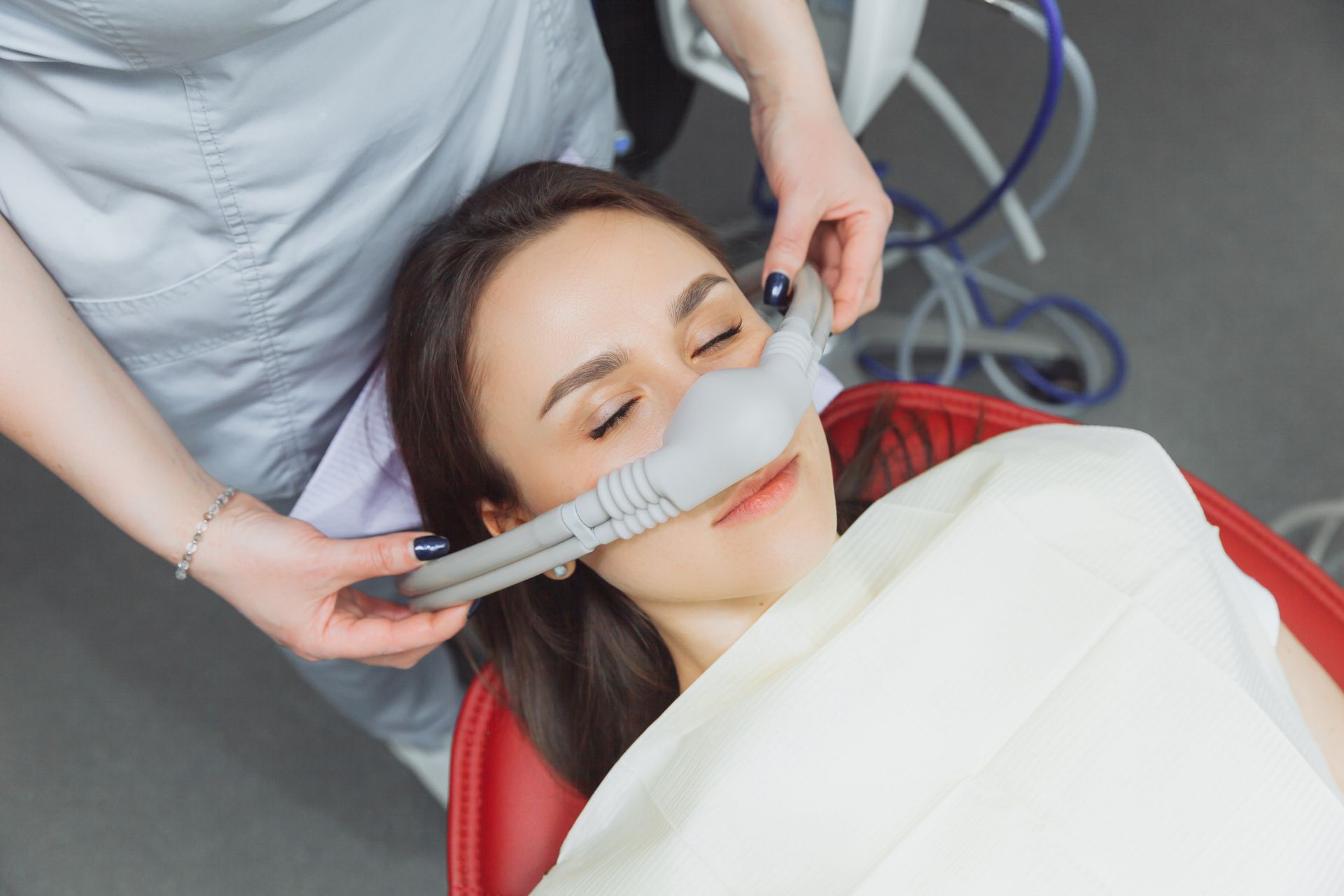 A dental assistant placing a mask on a woman's face in a dentist's chair; the woman has her eyes closed.