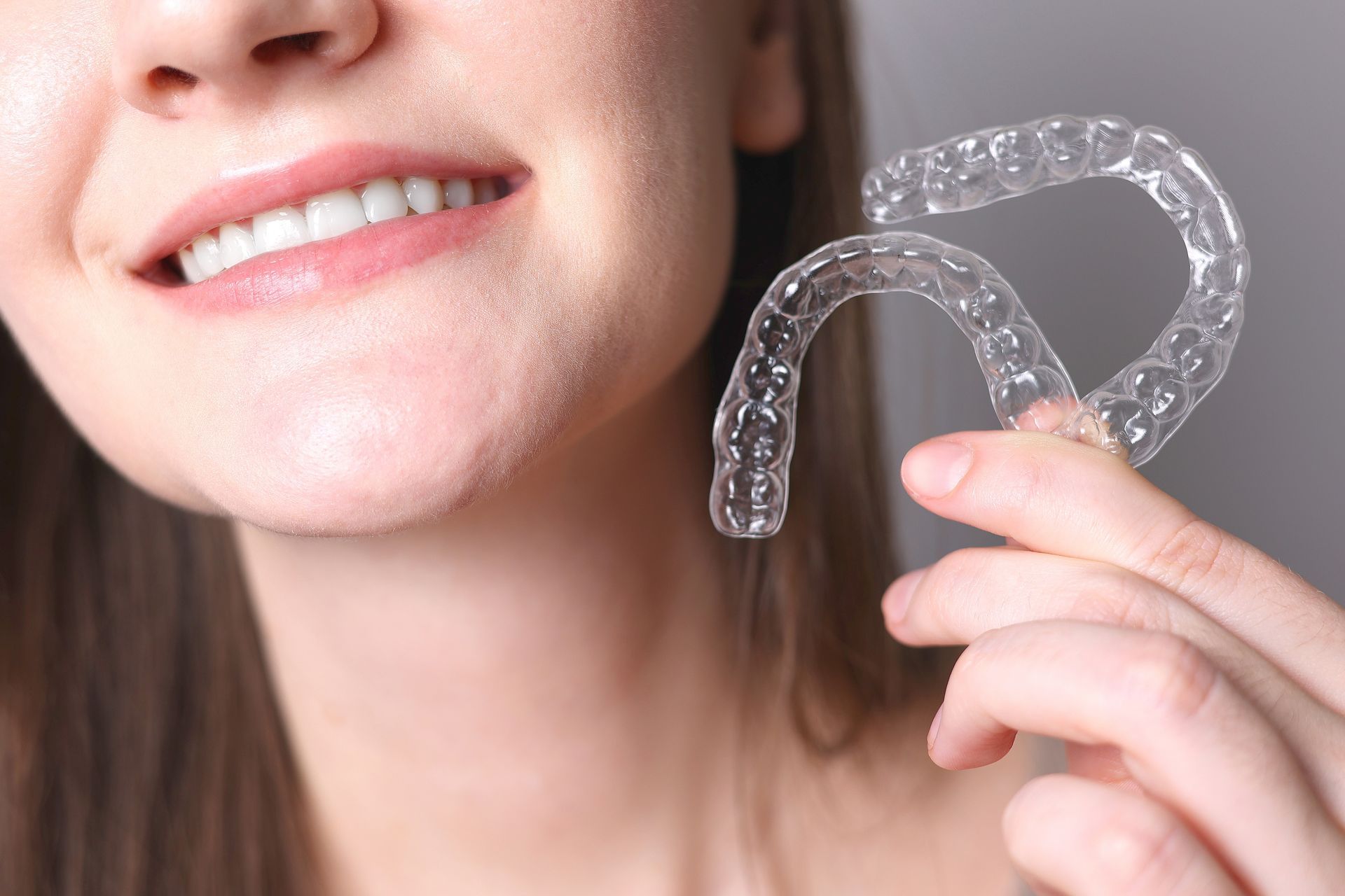 Woman holding clear aligners, smiling to show straight teeth; close-up shot against a neutral background.