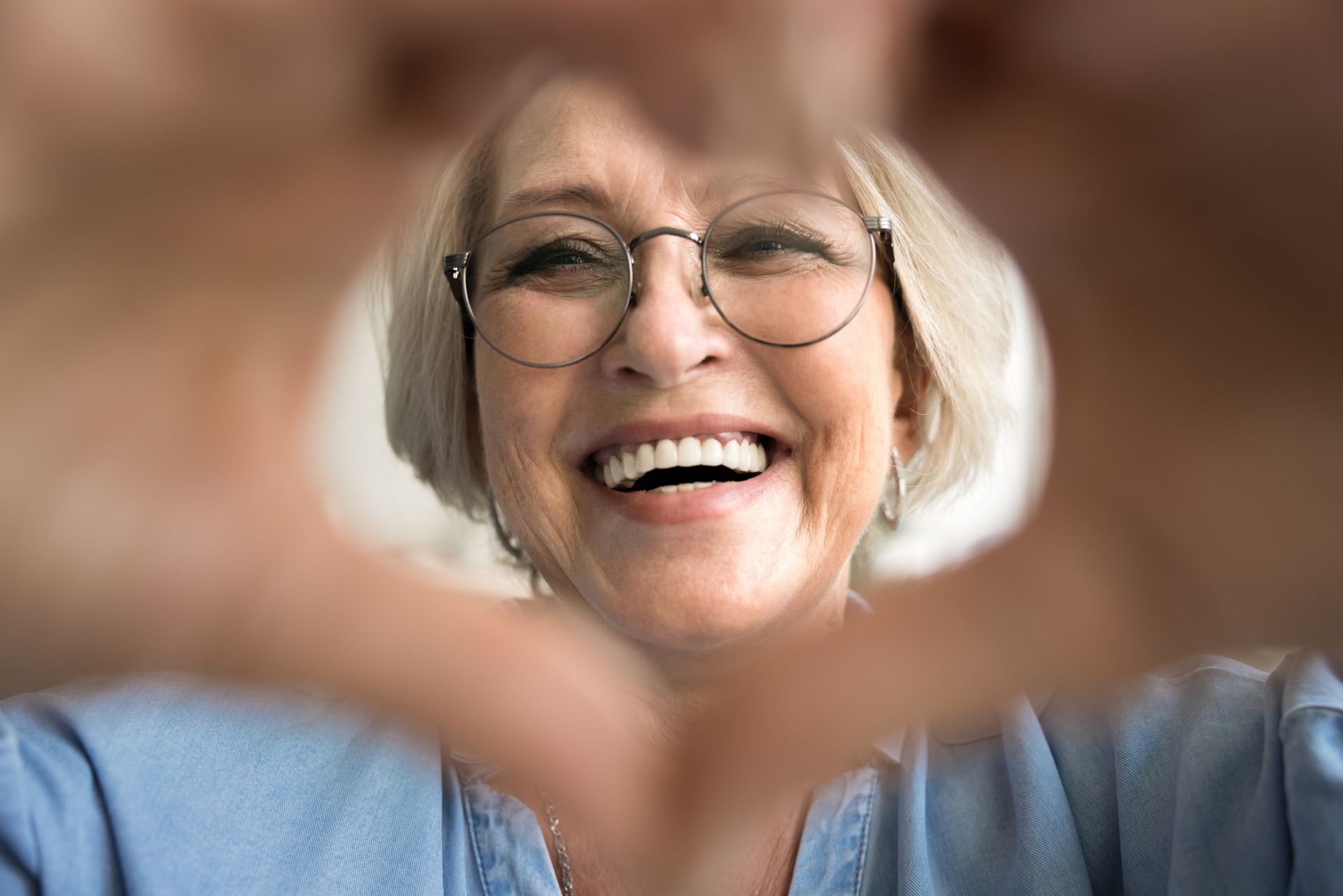 Smiling older woman wearing glasses forms a heart shape with her hands, blue shirt, indoors.