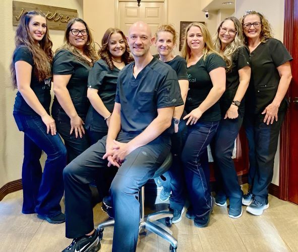A group of eight dental staff members pose in a dental office. A seated bald man in black scrubs is surrounded by seven women, all wearing black scrubs and dark jeans.