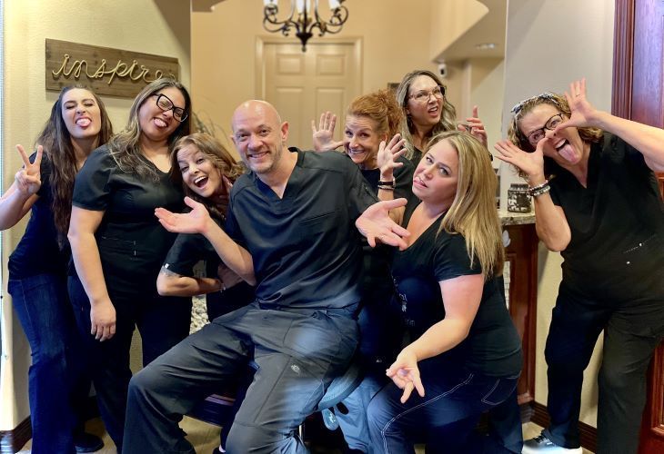 Group of dental staff smiling, making funny faces, and posing together in an office hallway. They wear black scrubs.