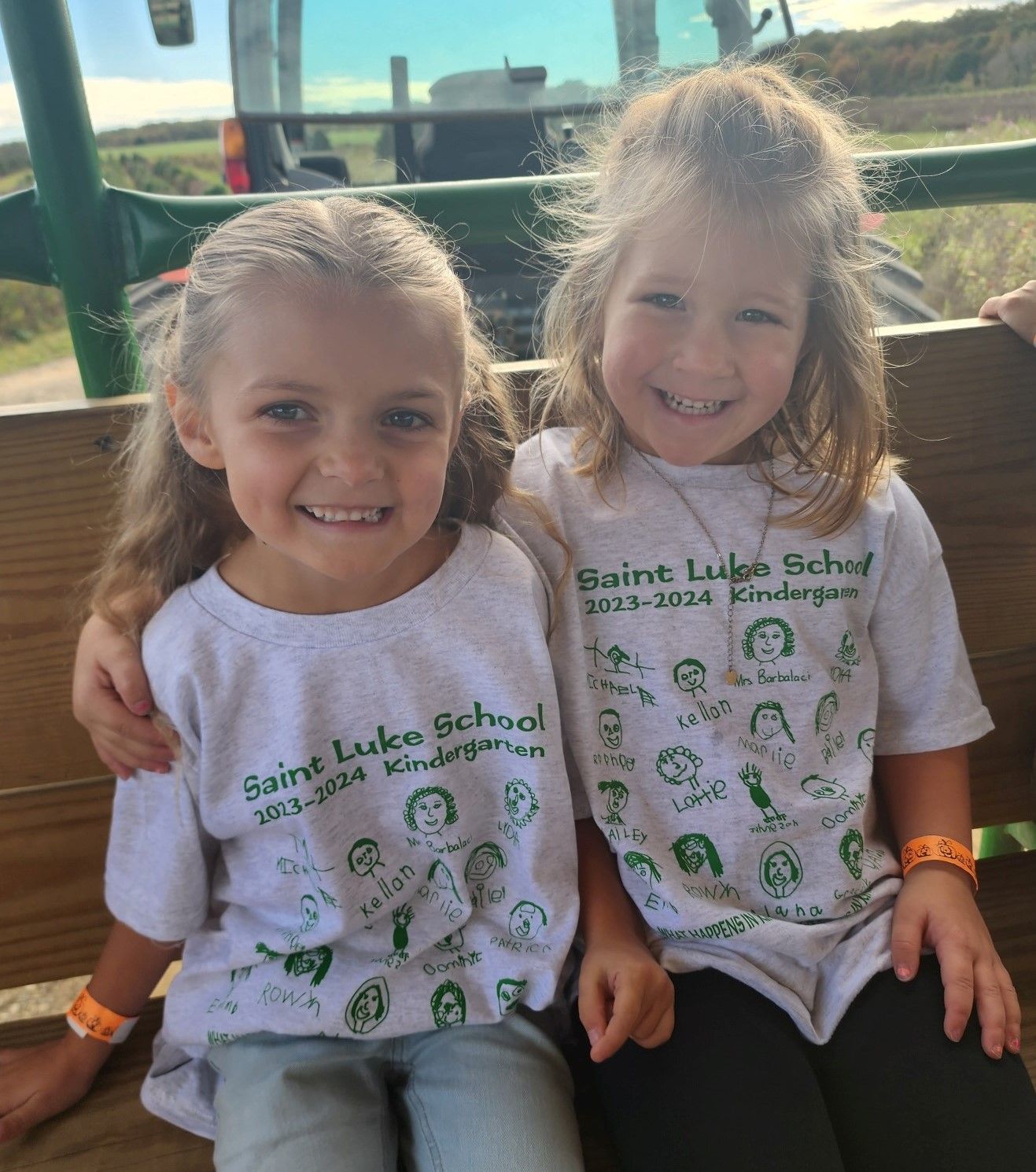 Two little girls wearing saint luke school shirts are sitting on a bench