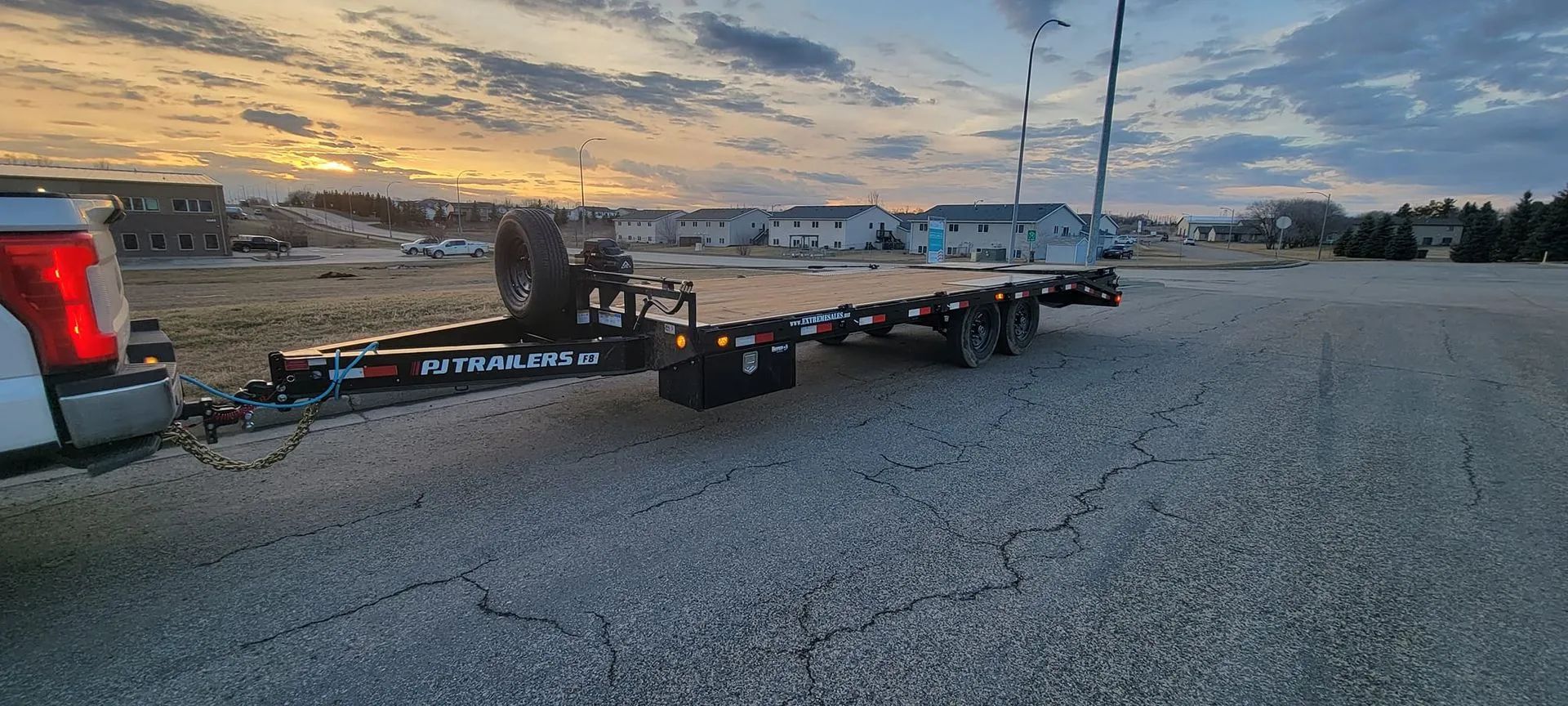 A truck towing a flatbed trailer on a cracked asphalt road at dusk with a residential neighborhood in the background.