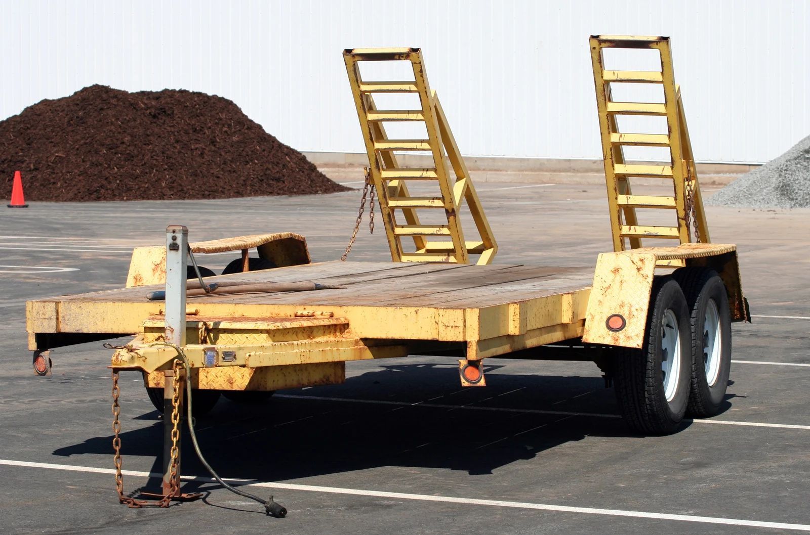Yellow flatbed trailer with ramps, parked on asphalt. Piles of mulch and gravel in the background.