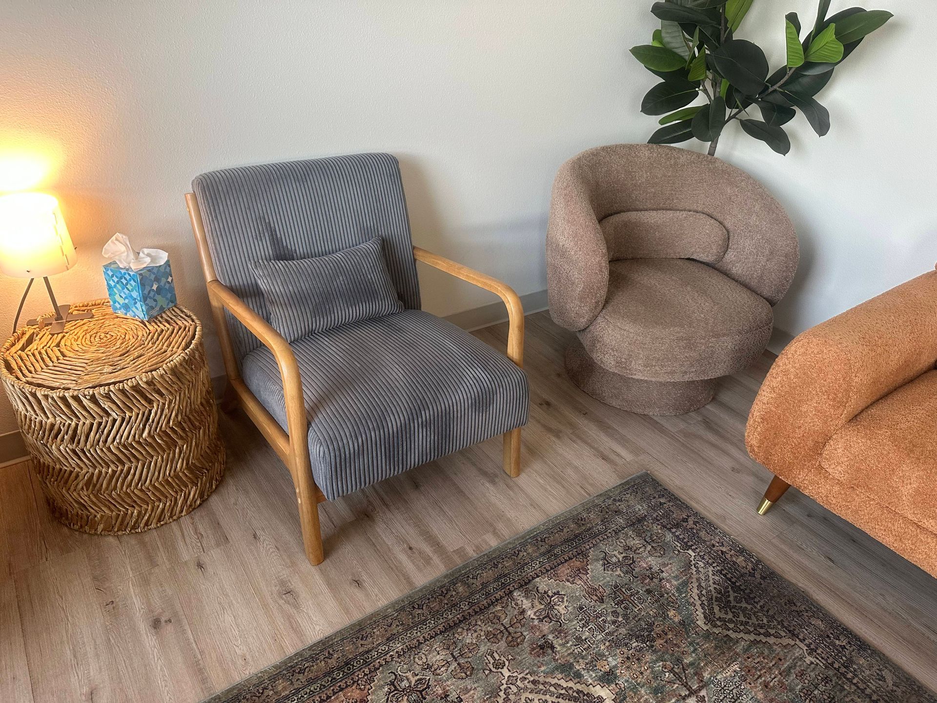 A cozy seating area with three chairs and a woven side table with a lamp and tissue box, on a rug.