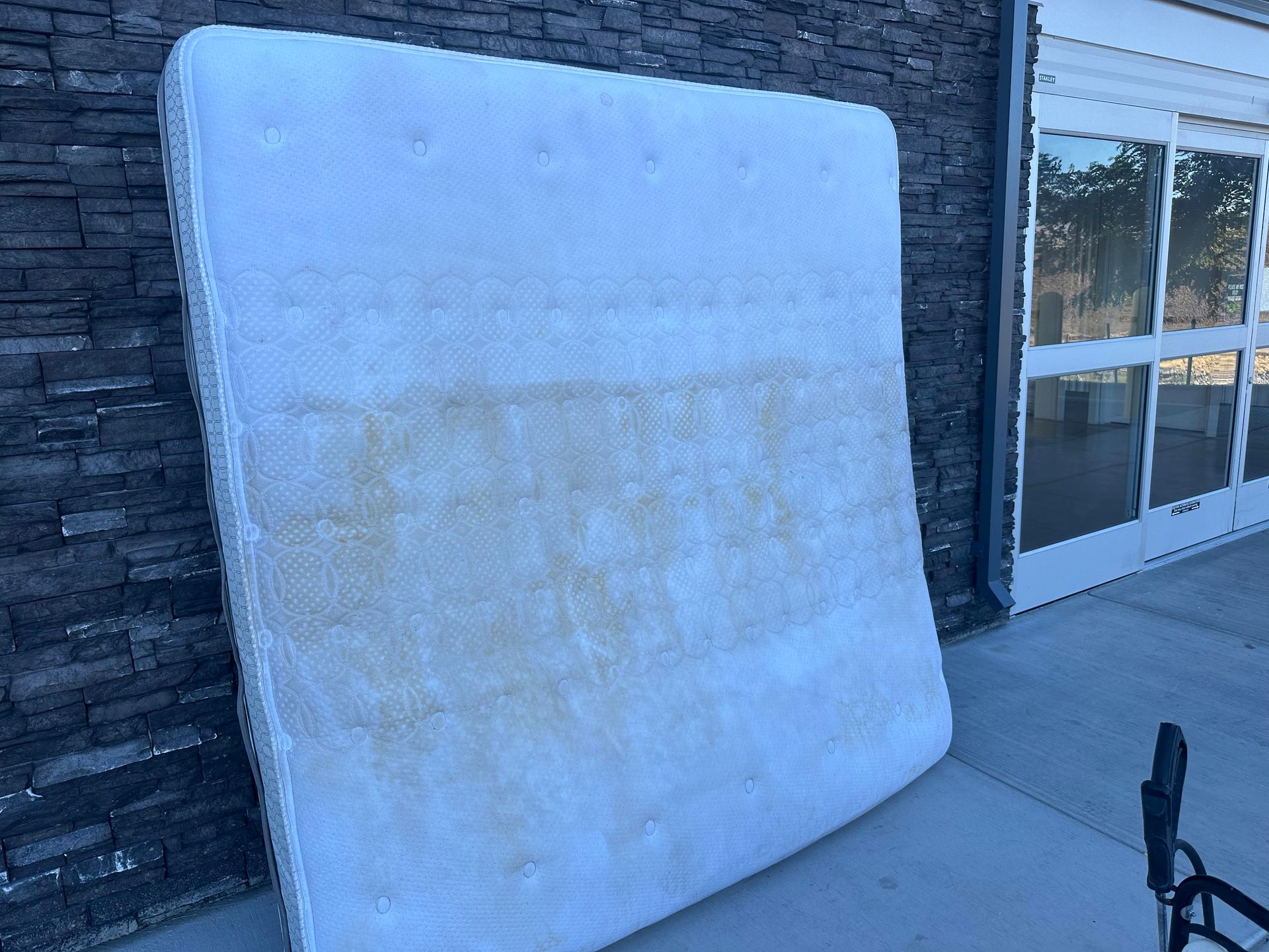 A stained mattress leans against a brick wall next to glass doors.