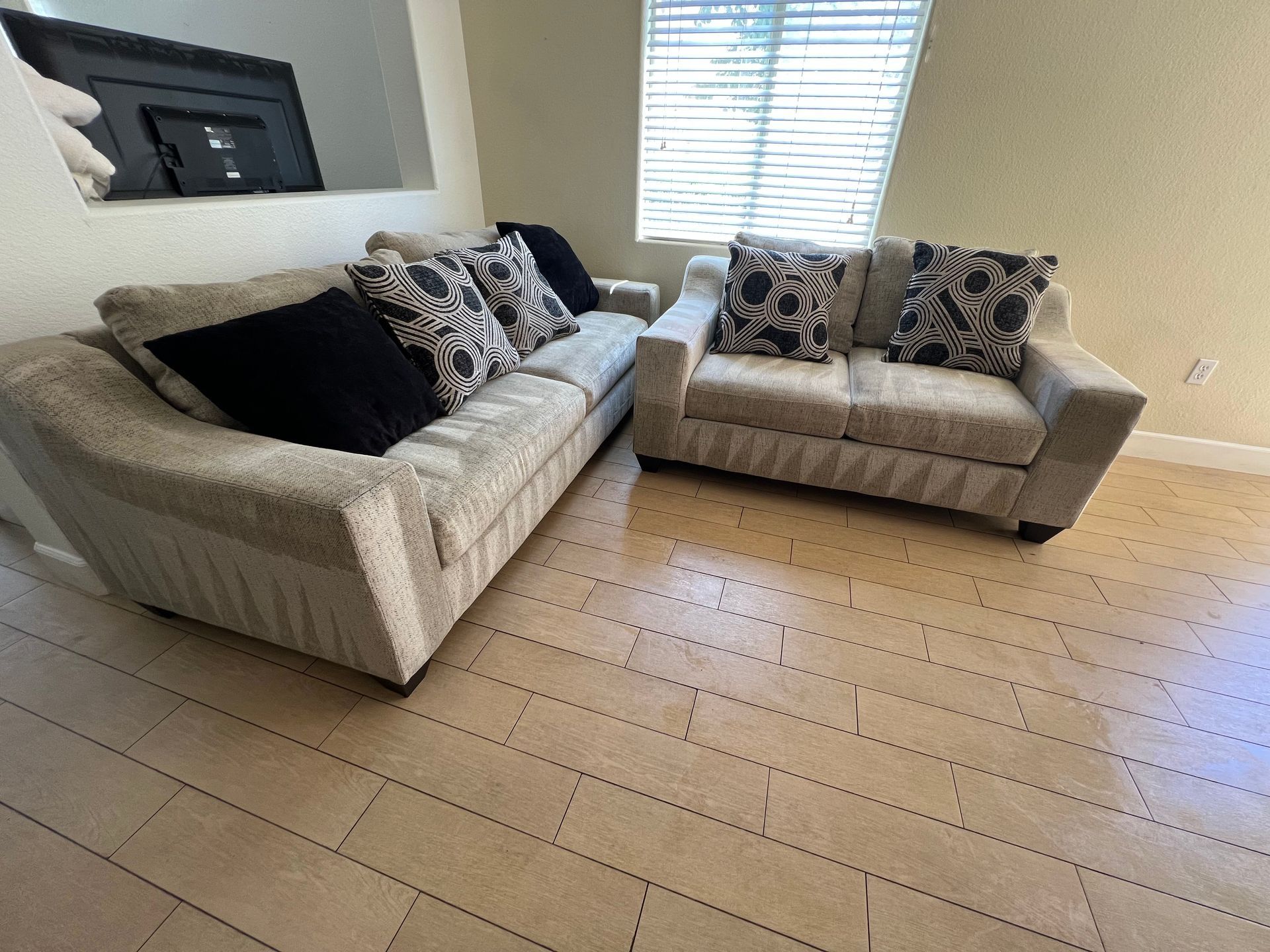 Two light-colored couches with patterned and black pillows, in a room with a window and tiled floor.