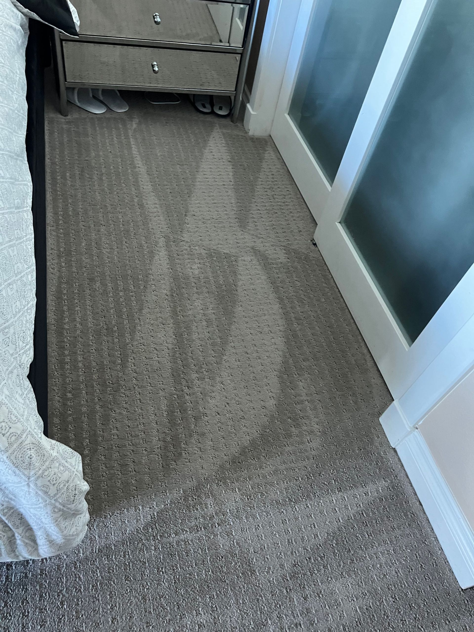 carpet with a noticeable lighter area, possibly from a bed or furniture, next to a mirrored nightstand and a glass door.