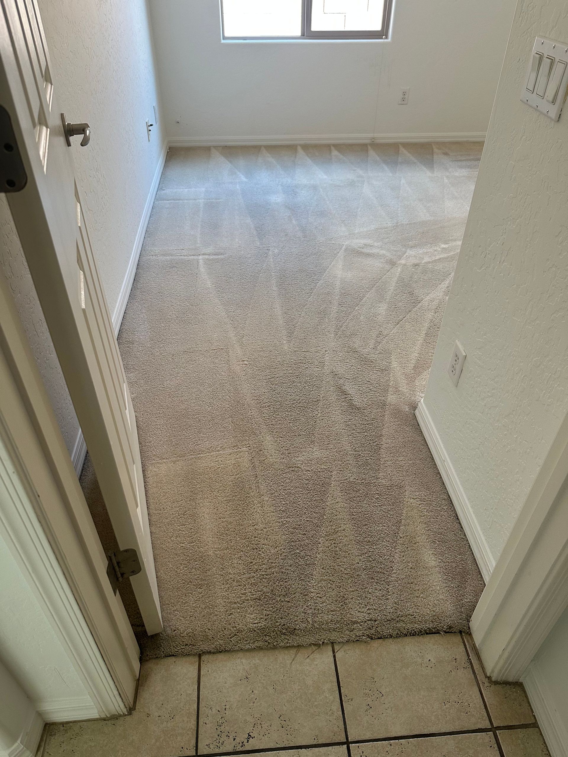 View from doorway into a room with worn beige carpet, a window, and white walls.