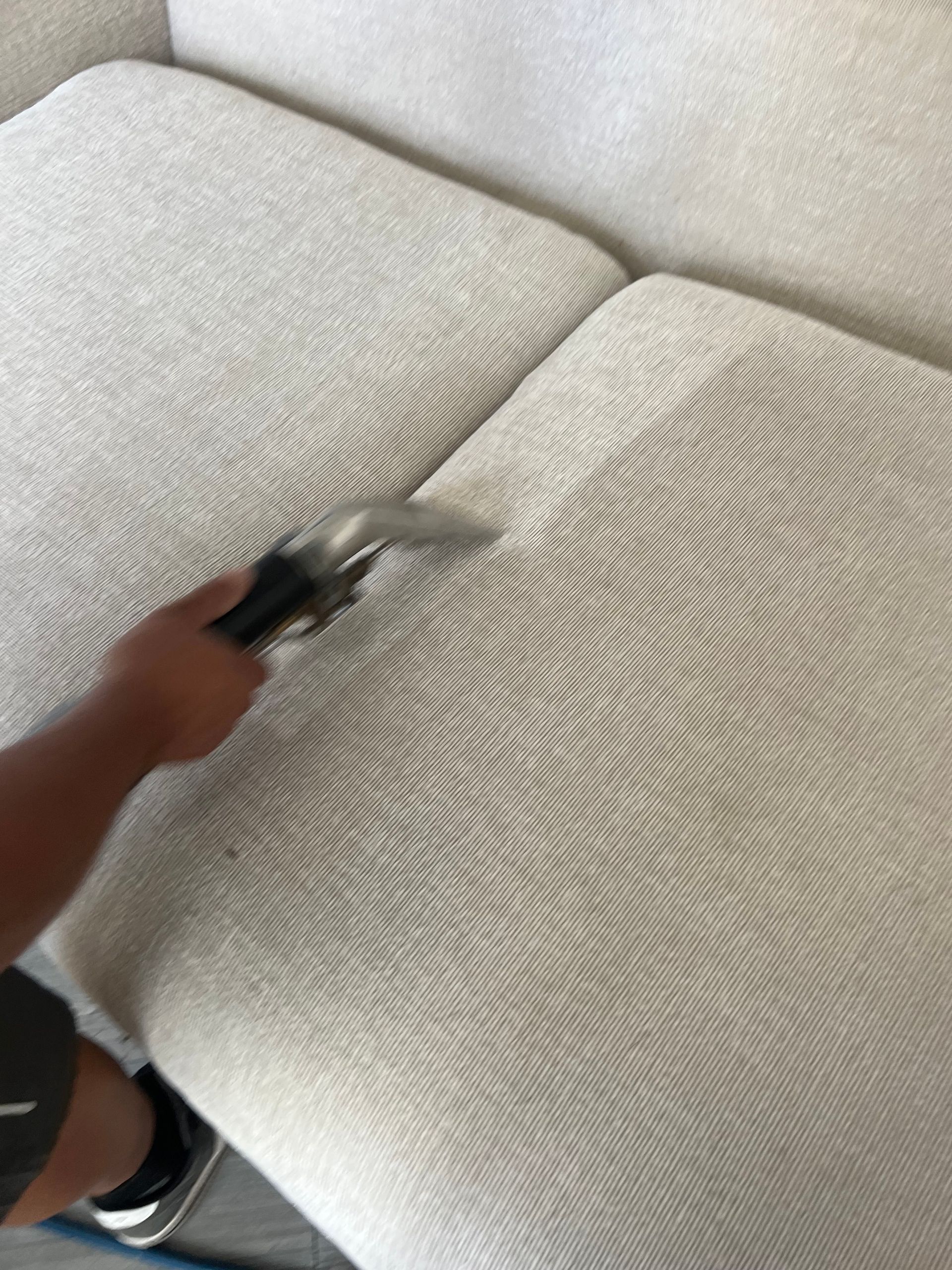 Person cleaning a light-colored sofa with a handheld upholstery cleaner.