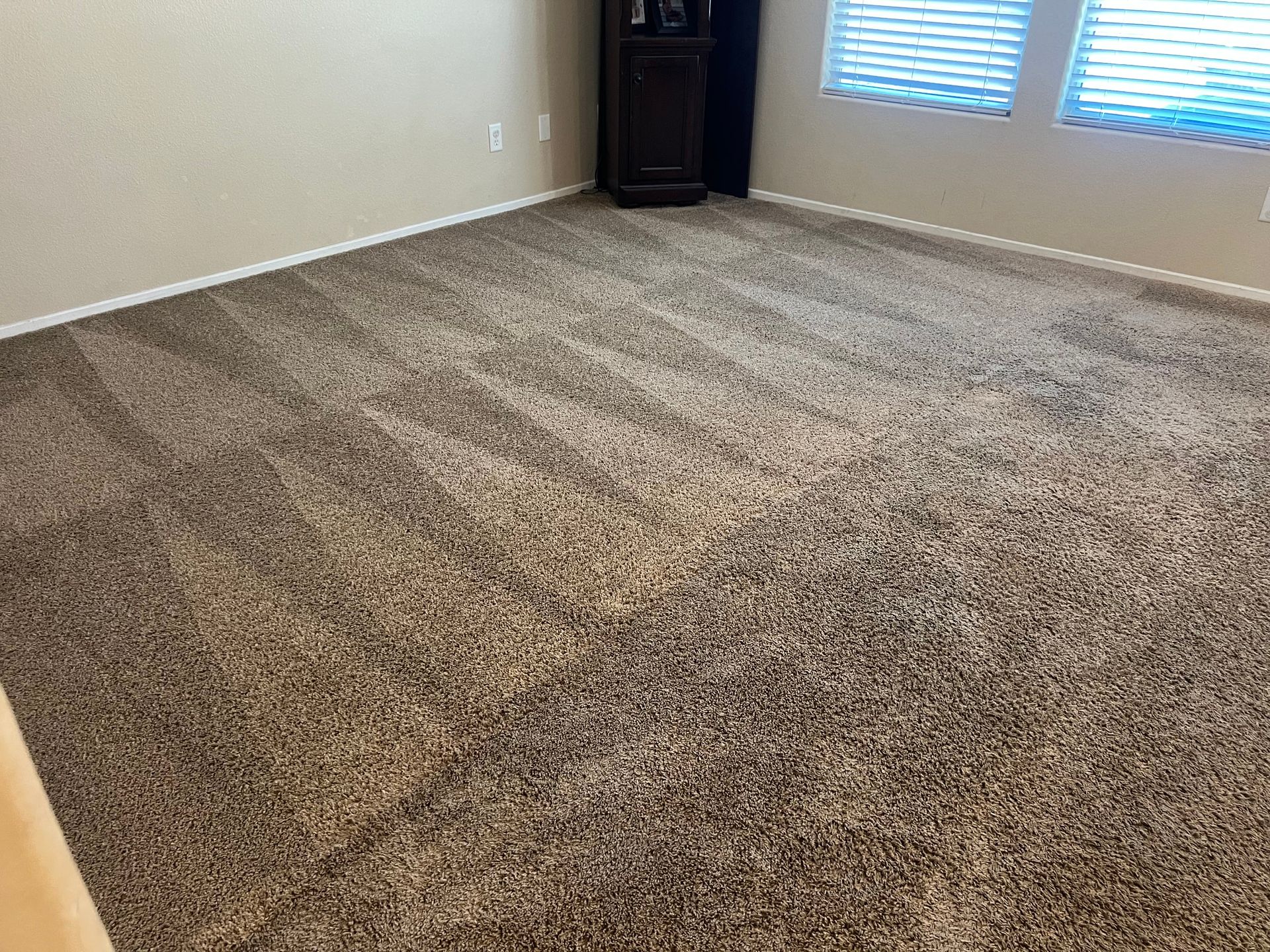 Brown carpet in a room with a corner cabinet and windows with blinds.