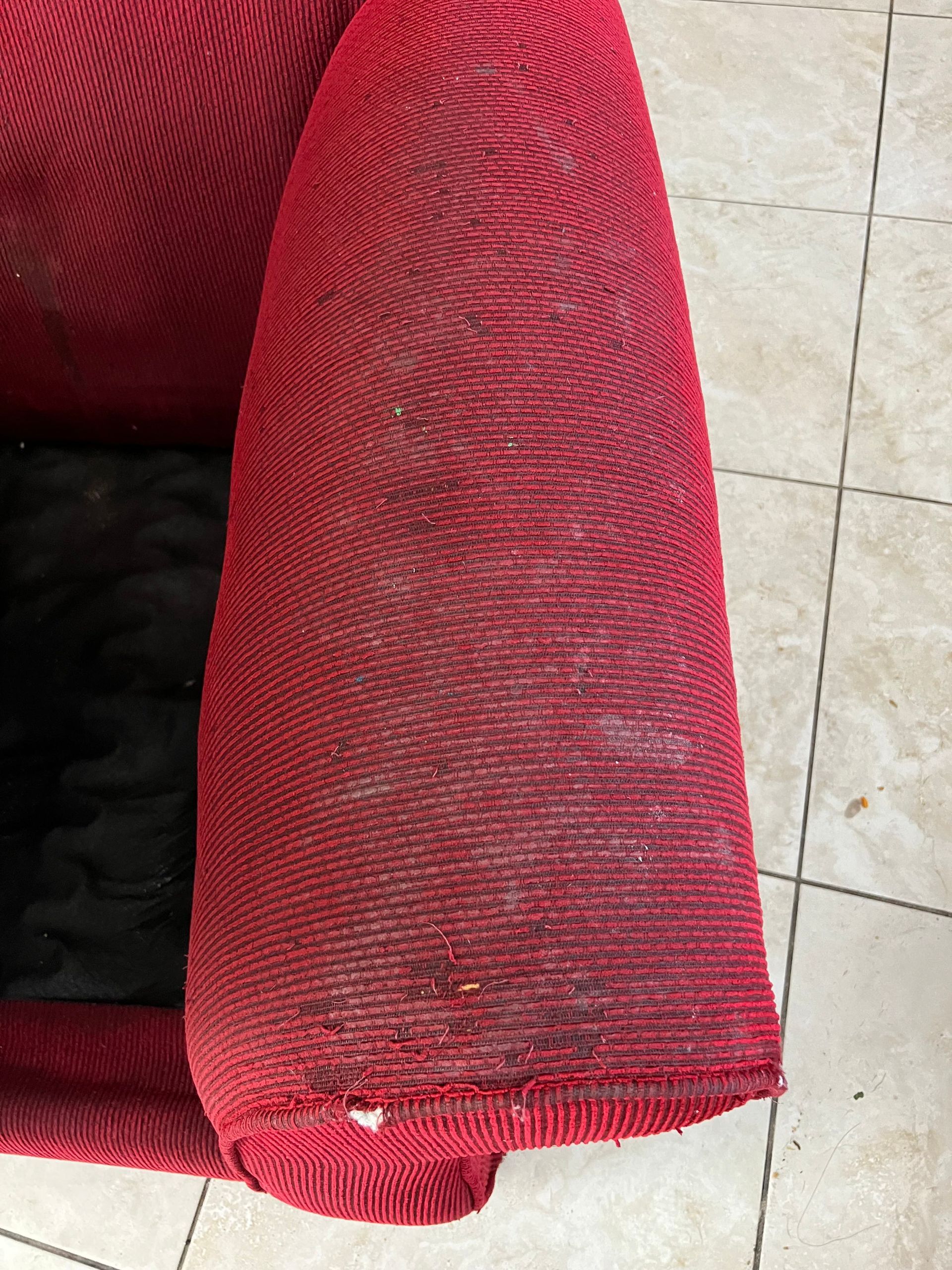 Red fabric armchair armrest, showing woven texture and wear, on tiled floor.