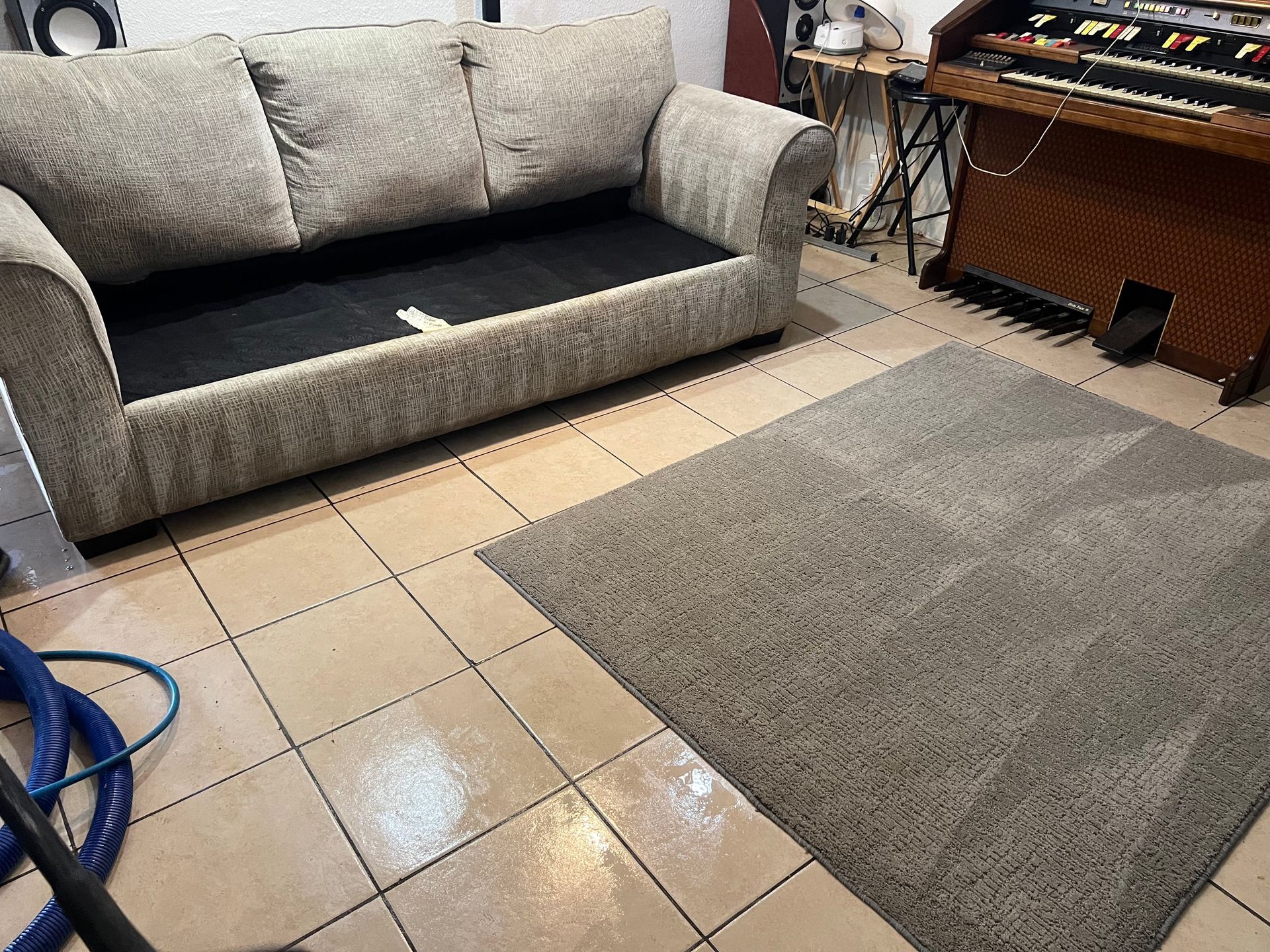 A light-colored sofa with a damaged cushion sits on tiled floor, near a rug and musical equipment.