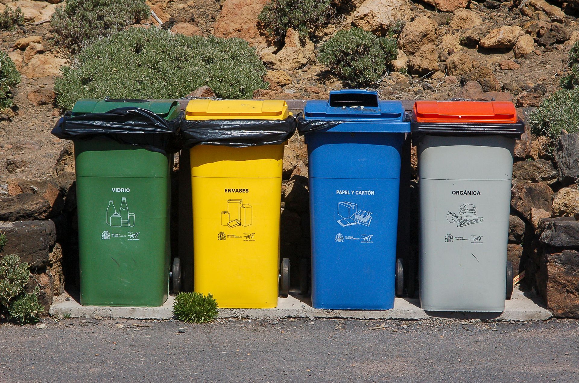 Four colorful recycling bins: green, yellow, blue, and gray, with corresponding lids, against a stone wall.