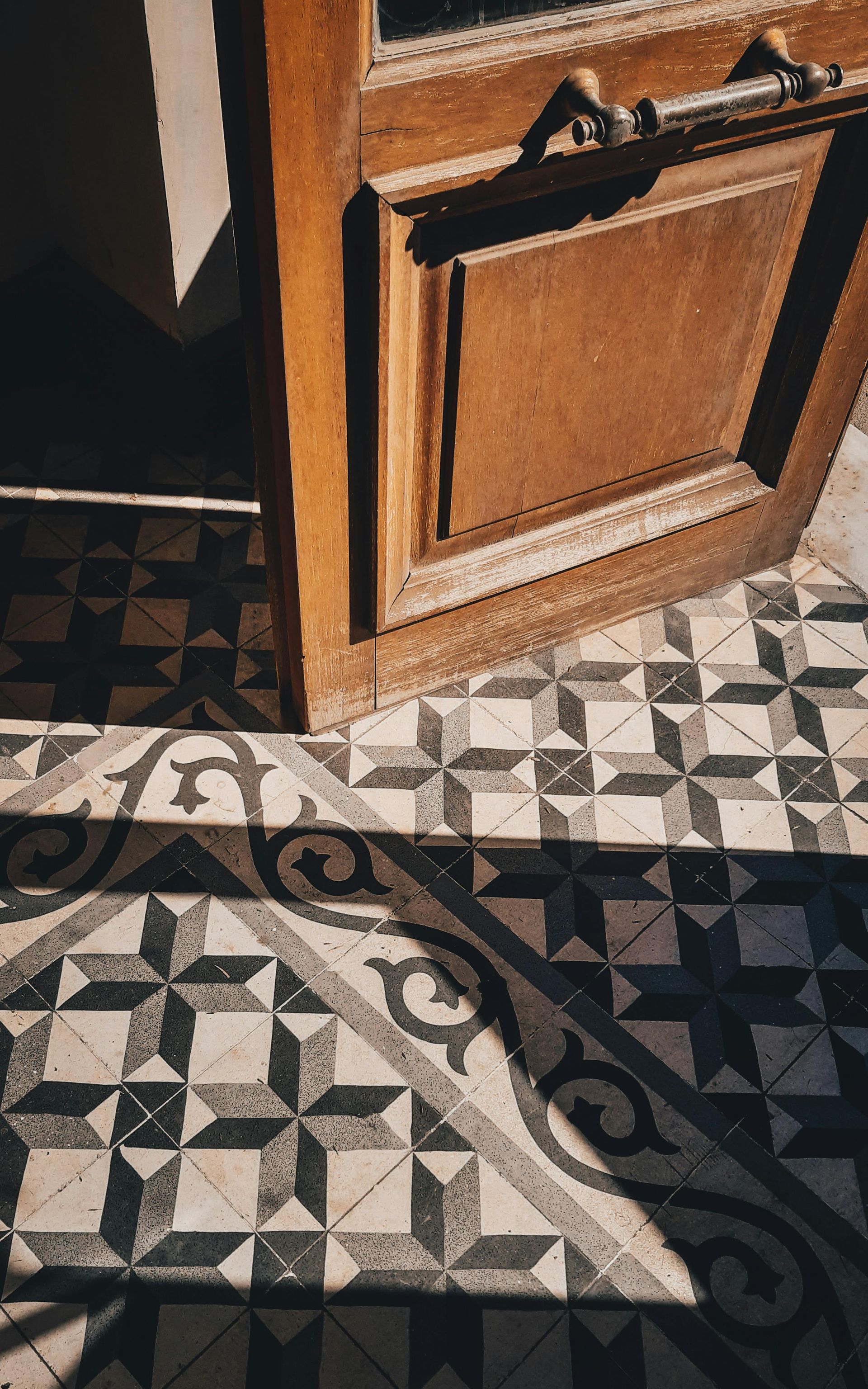 Wooden door slightly ajar over a black and white patterned tile floor, casting a shadow.