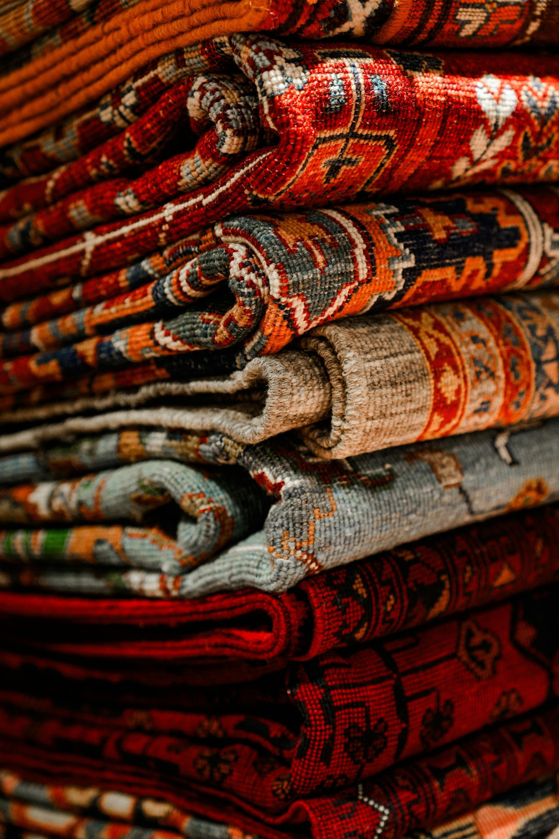 Pile of colorful, patterned rugs stacked on top of each other. Various shades of red, orange, and blue.