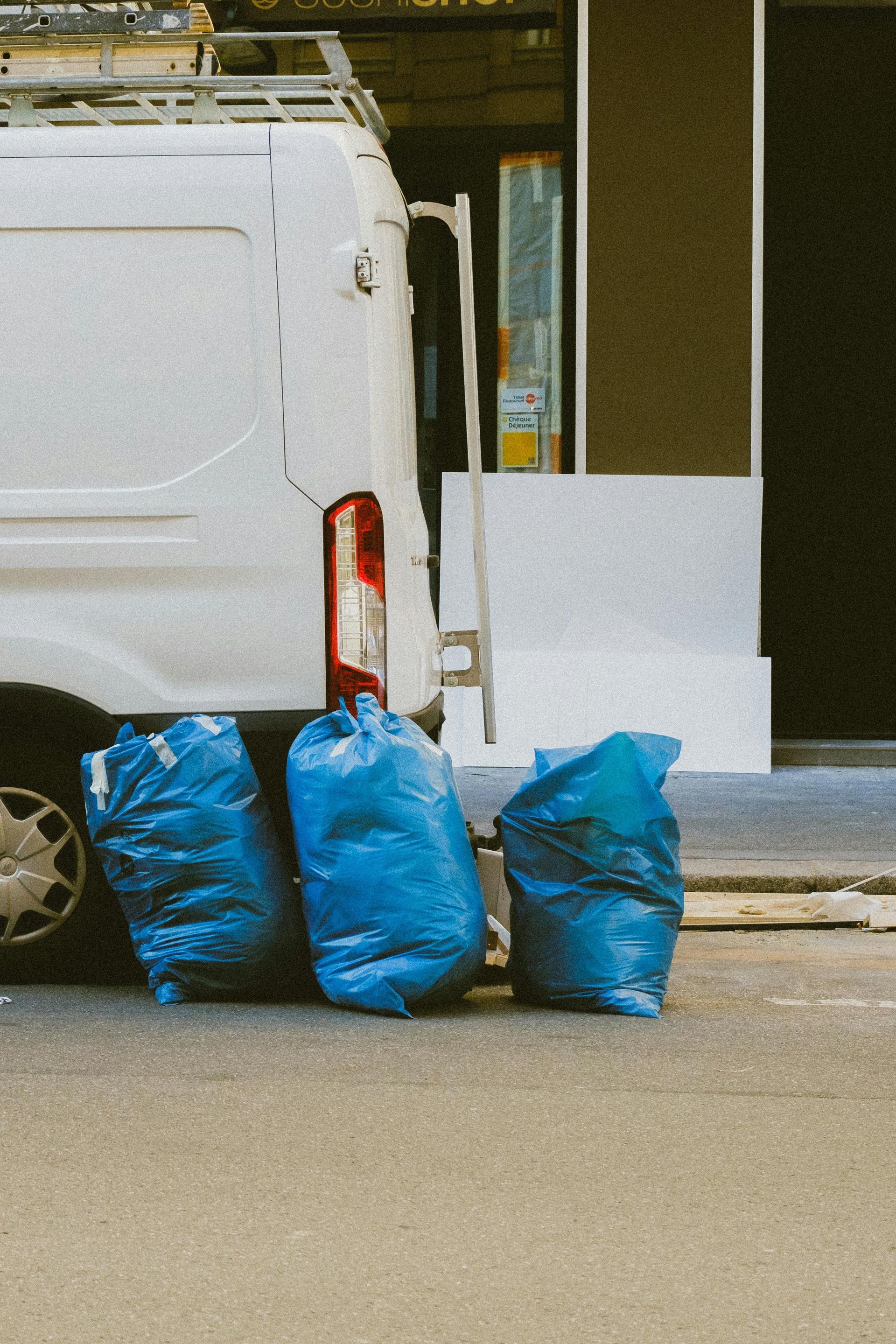 Three blue trash bags on a city street next to a white van and a building.