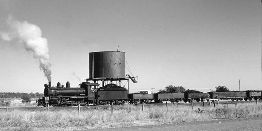Black and white photo of a steam train pulling coal cars next to a water tower.