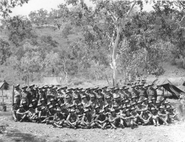 Group of soldiers in uniform posing outdoors, tents and trees in the background.