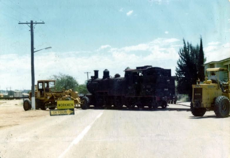Steam engine being moved on a road, flanked by road graders. Warning sign in foreground.