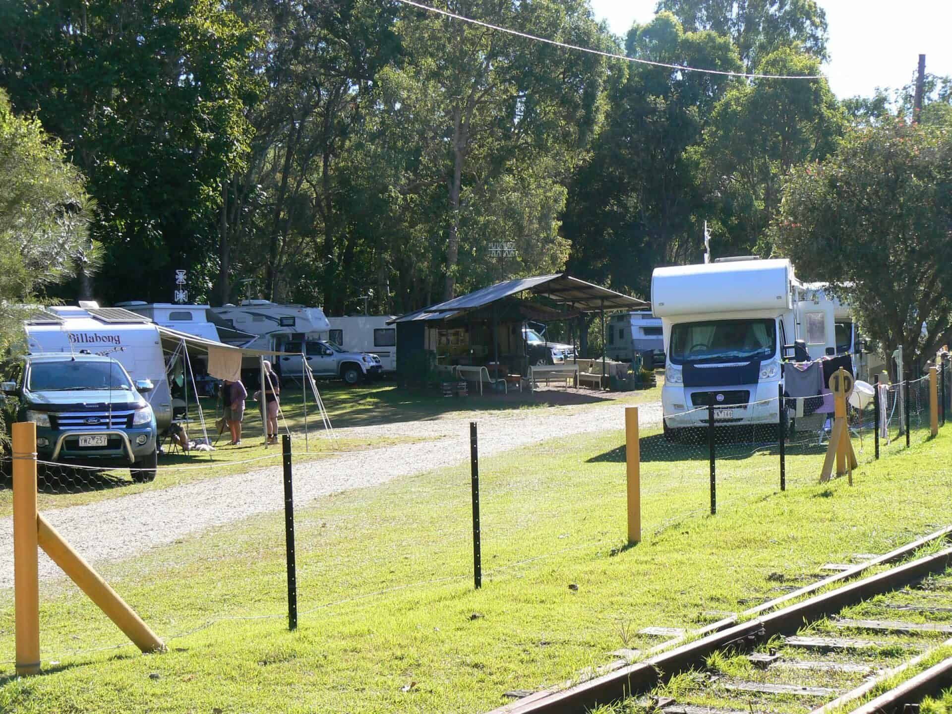 Campground with RVs, cars, and a shelter next to train tracks, surrounded by trees.