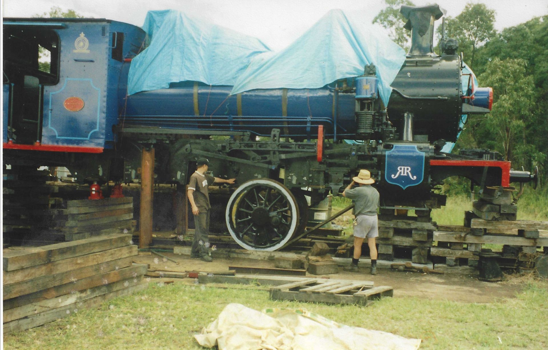 Steam locomotive on blocks, covered with tarp, being worked on by two men outdoors.