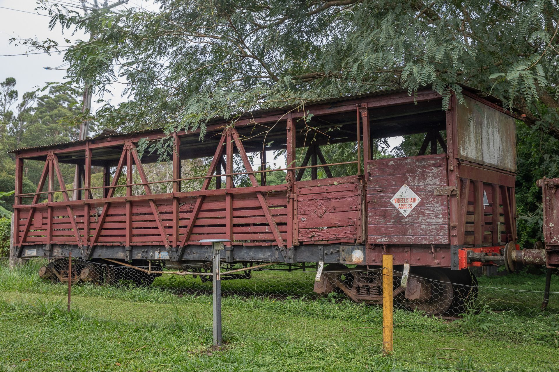 Old, weathered red train car, open-sided with wooden slats, resting in grassy area by trees.