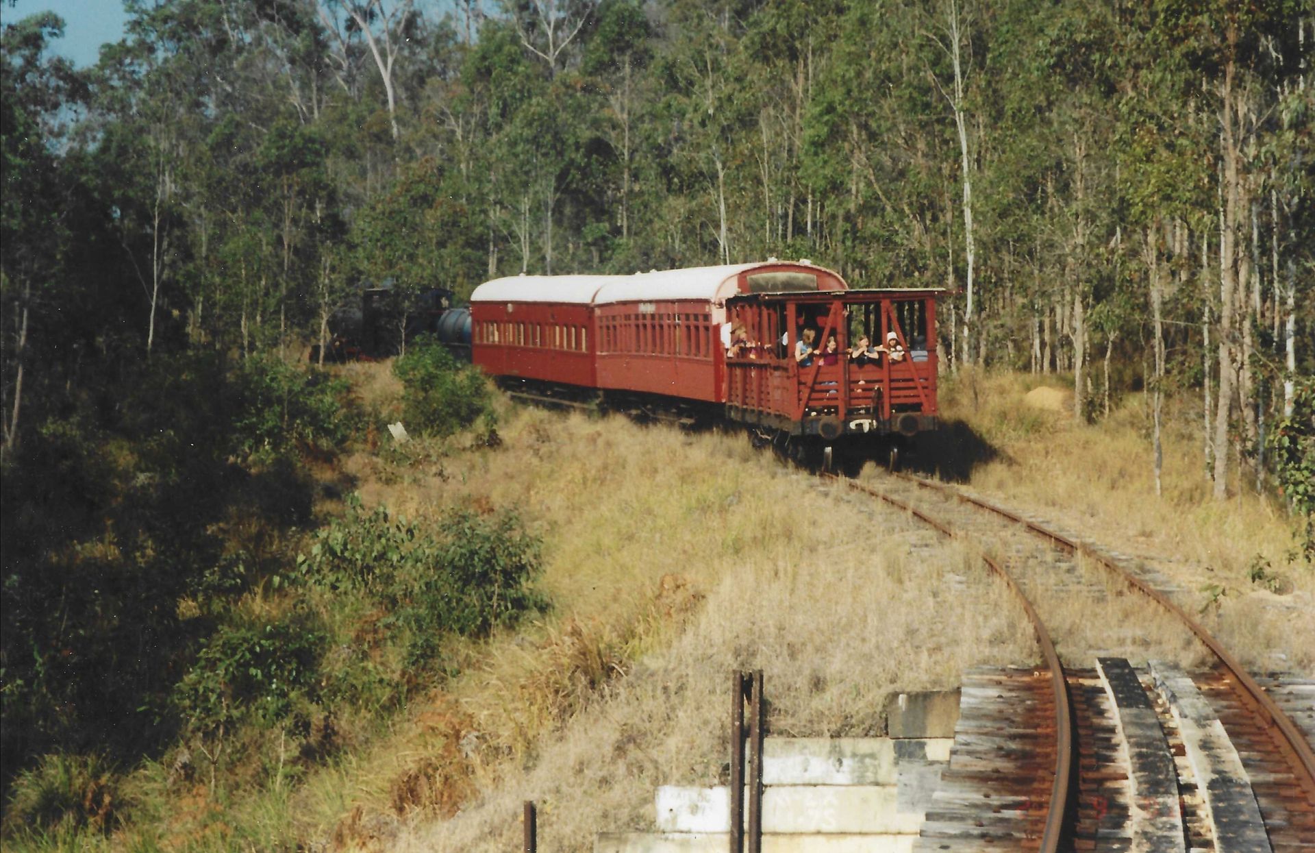 Red train on tracks in a wooded area, with people visible.