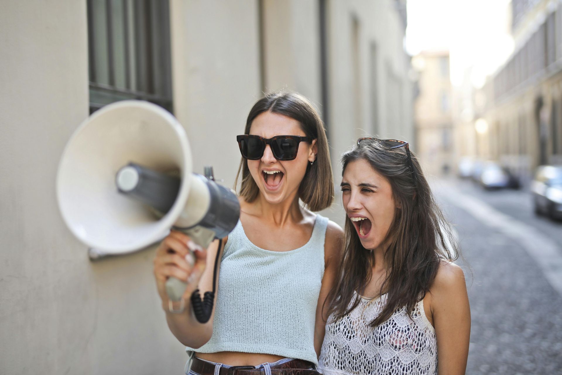 Two women shouting into a megaphone on a city street. One wears sunglasses.