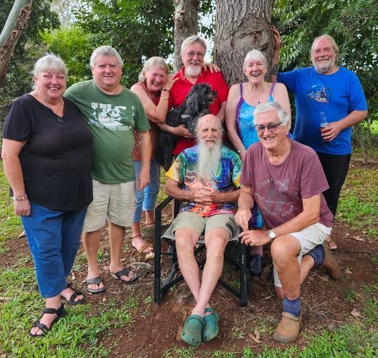 Group of people smiling outdoors, gathered around a seated man with a long beard.
