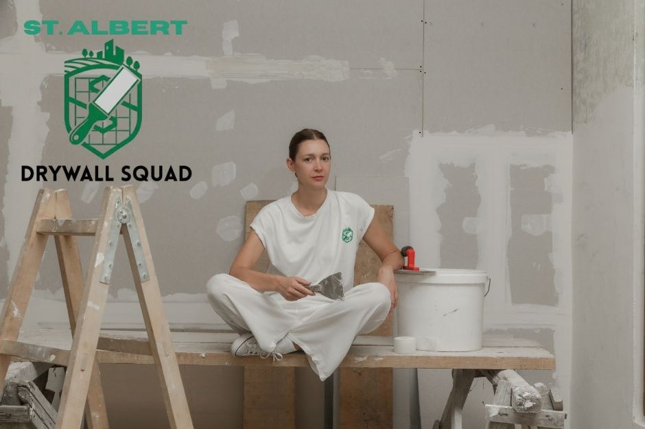 A drywall repair company worker sits on wooden planks during a commercial renovation, wearing white work clothes. She holds a putty knife while taking a break, with a stepladder, paint bucket, and construction materials visible around the unfinished room near Huron Dr, and Athapaskan Dr, Leduc, AB.