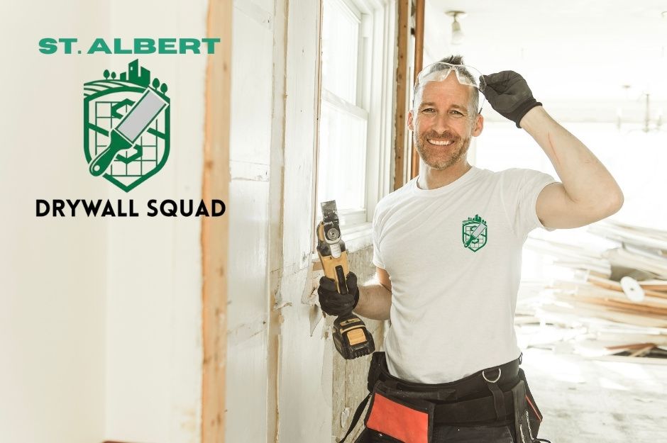 A drywall repair company worker smiles while holding a power drill at a residential construction site with exposed wooden framing and white walls. He wears work clothes and safety glasses, near Summercourt Rd, and Summerwood Blvd, Sherwood Park, AB.