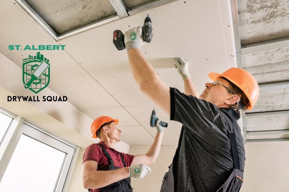 Drywall repair contractors work on ceiling installation. Two workers in orange hard hats and work clothes use cordless drills to secure drywall panels to the metal ceiling framework inside a commercial construction site near Graywood Terrace, and Graybriar Dr, Stony Plain, AB.