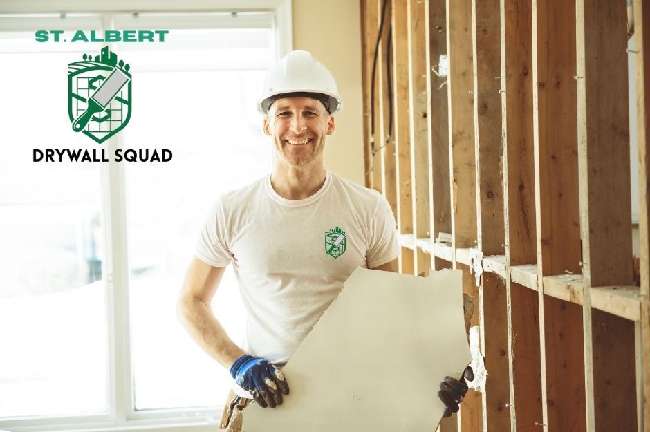 A drywall repair contractor in a white hard hat and shirt smiles while holding a drywall sheet against wooden wall studs in a bright commercial construction site. A professional contractor prepares for installation work near 49 Ave, and 40 St, Beaumont, AB.