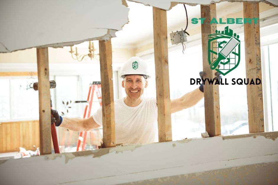 A drywall repair specialist in a white hard hat smiles while holding a sledgehammer during an interior renovation project, standing among exposed wooden ceiling joists and damaged drywall in a residential space located near Rancher Rd, and Horton Wy, Ardrossan, AB.