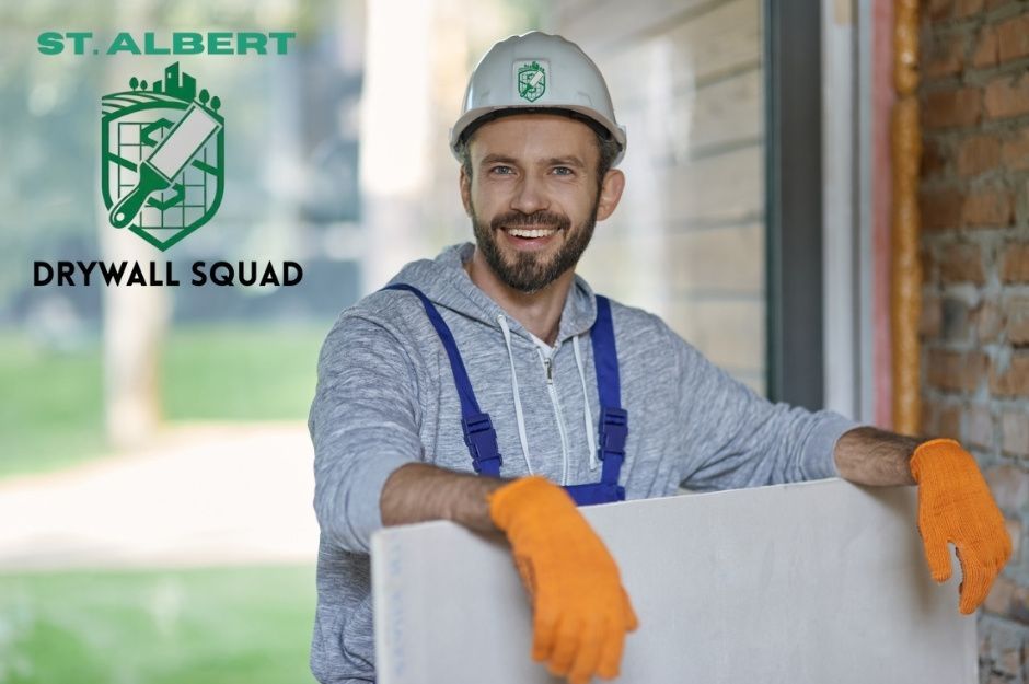 A drywall repair technician in a gray hoodie and white hard hat smiles, wearing orange work gloves and leaning against white drywall sheets at a construction site. He wears blue overalls in a commercial setting near Range Rd 244A, and 50302 Range Rd 244A, Nisku, AB.