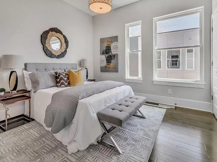 A bedroom with a bed , a bench , a mirror and two windows.