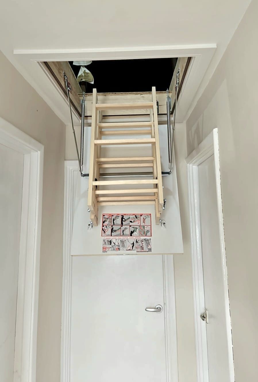 Hallway with pull-down attic stairs. Wooden ladder extends from an open ceiling hatch. White door on the lower level.
