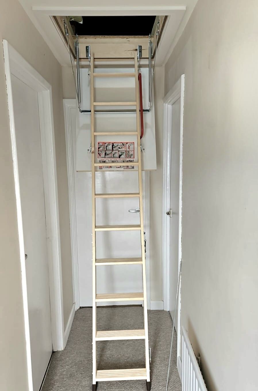 Attic ladder extended from ceiling access in a hallway. Beige walls, white doors, and a ladder made of light-colored wood.