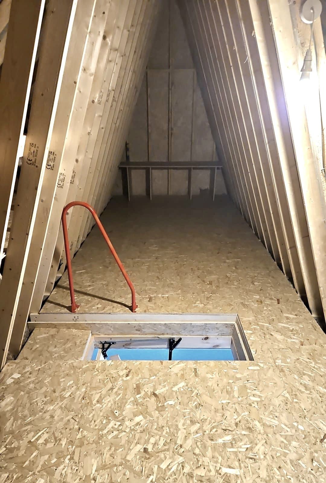 Interior of an unfinished attic with plywood floor and roof, access hatch, and a red safety bar.