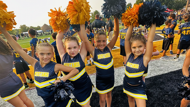 Flag football game at night, a player in red jersey runs with the ball as others pursue on a green field.