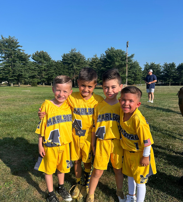 Flag football game at night, a player in red jersey runs with the ball as others pursue on a green field.